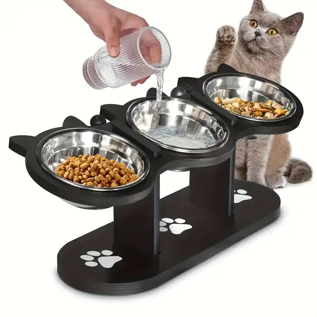 A stylish black cat-shaped feeding station with three elevated stainless steel bowls for food and water, as a person pours water into one of the bowls. A curious grey cat sits nearby, raising its paw playfully.