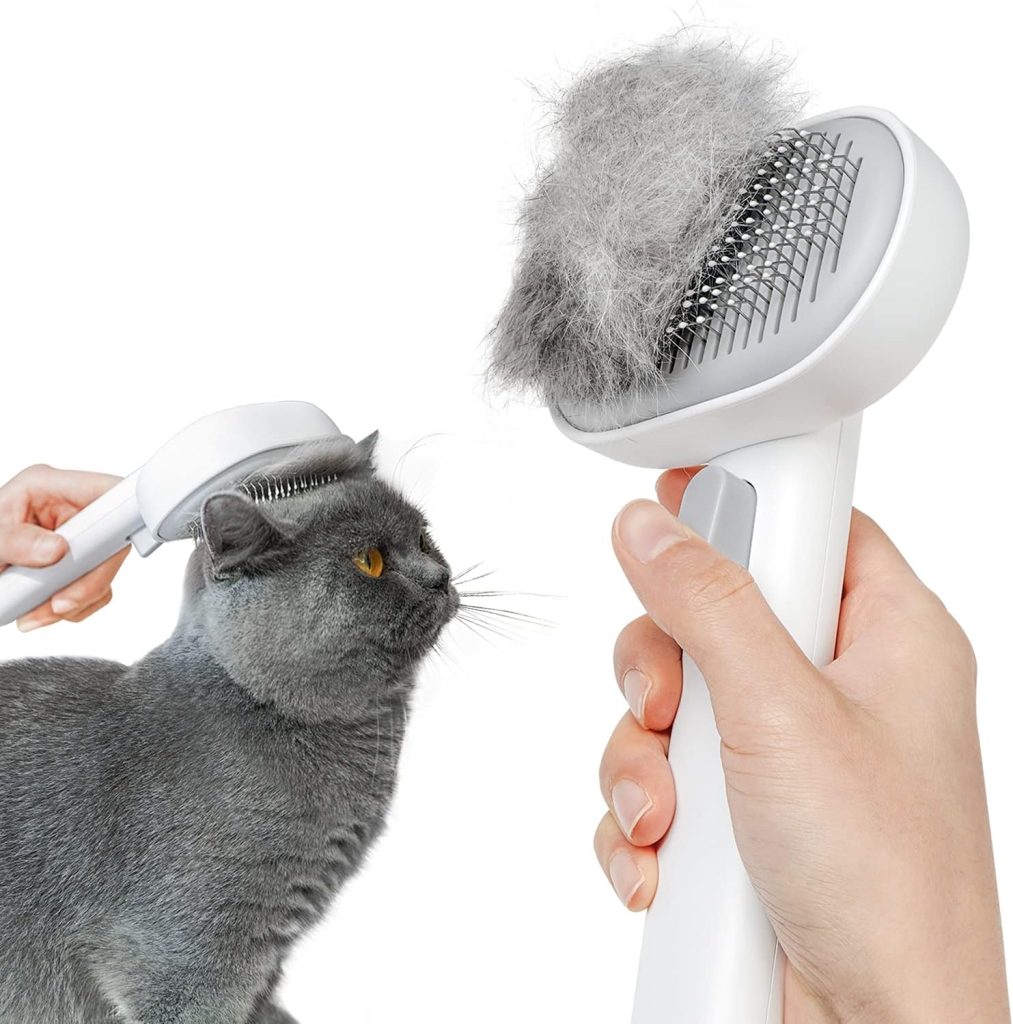 A grey cat being groomed with a white self-cleaning cat brush, shown with a hand pressing the brush's cleaning button to release collected fur.